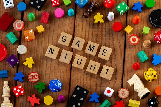 Scrabble letters spell out the words game night on a wooden tabletop the words are surrounded by a wide variety of game pieces in a rainbow of colors.