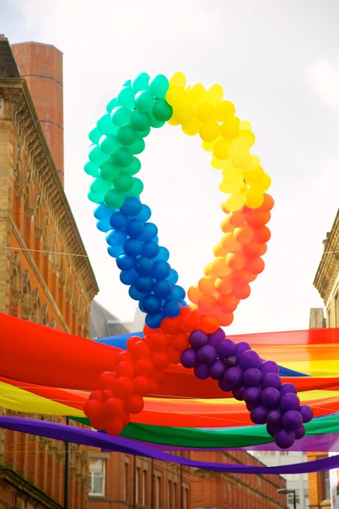 An arch of rainbow balloons hangs in the air between rows of buildings,; behind them rainbow streamers hang across the road.